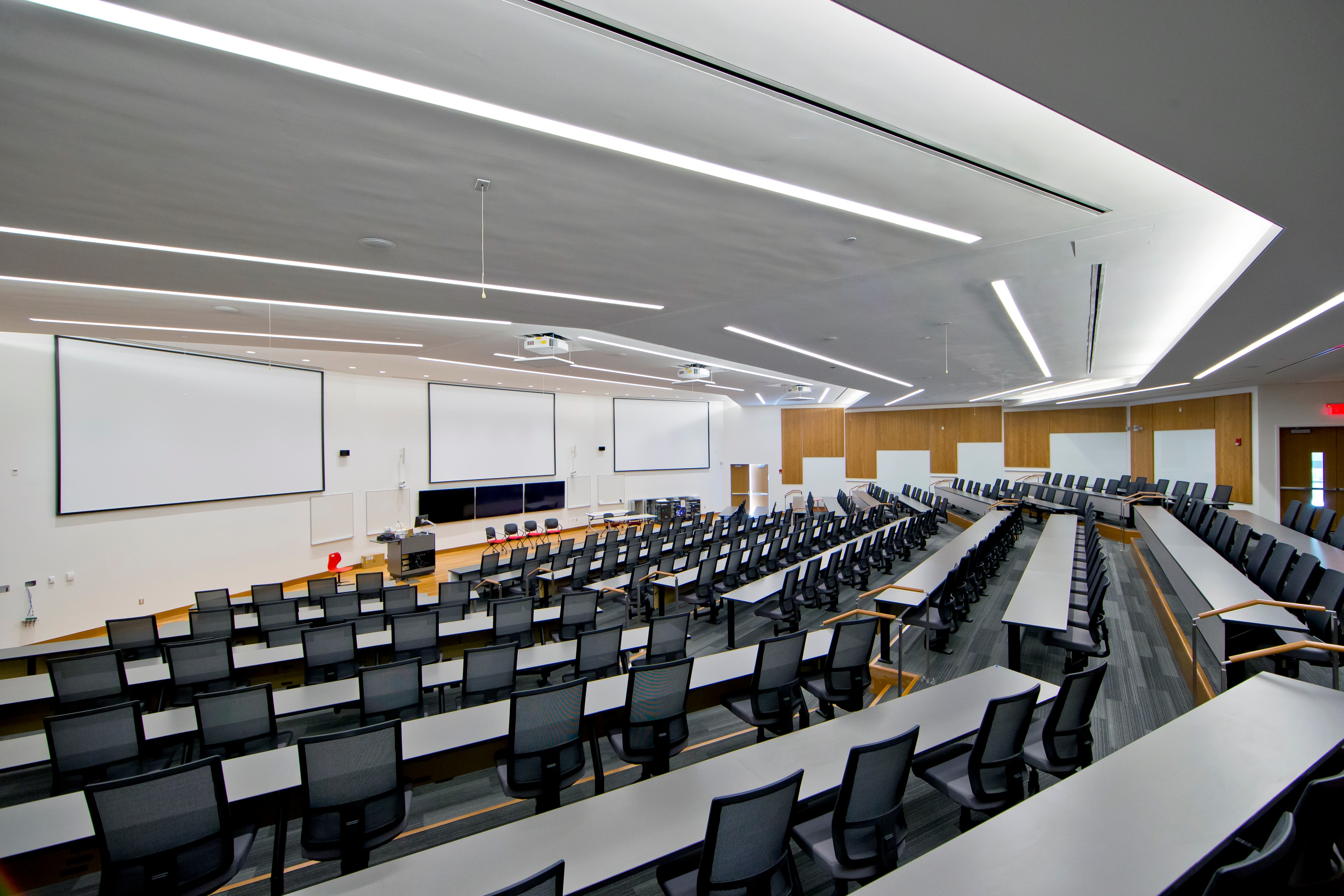 A large room with rows of long gray tables, each table with several black chairs positioned to face a wall of whiteboards.