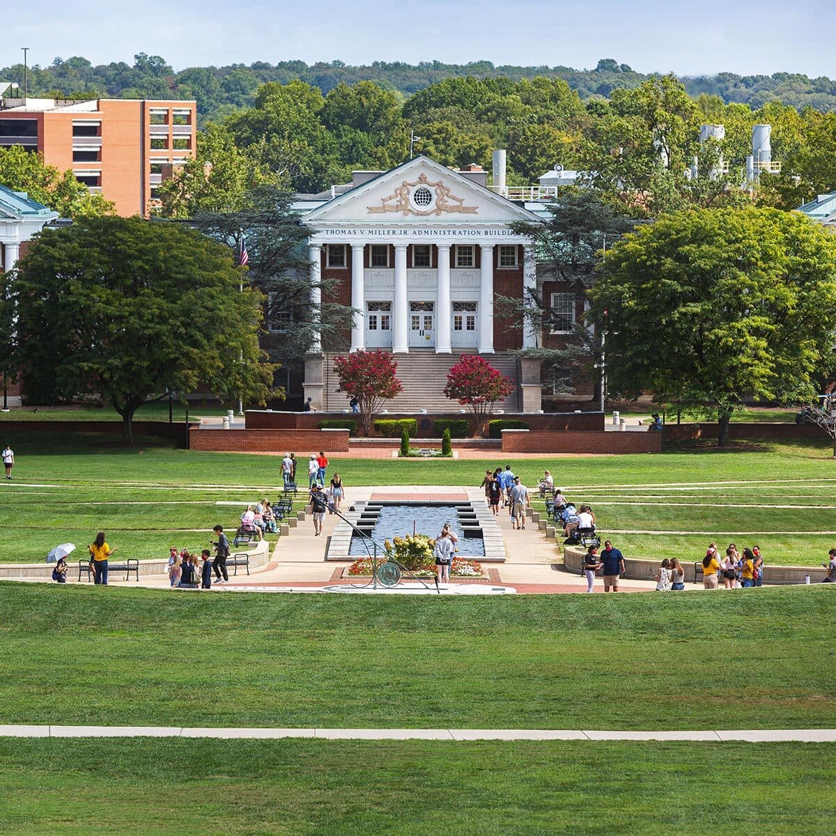 Wide view of the lush and green heart of the University of Maryland campus, McKeldin Mall. Community members walk along pathways, or mingle around the long gentle ODK Fountain